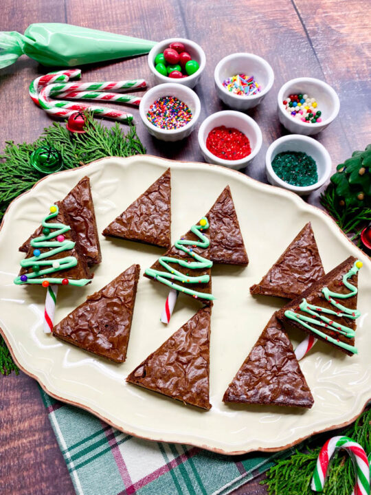 Christmas tree brownies on a plate with bowls with sprinkles, candy canes and a piping bag with icing for a decorating station