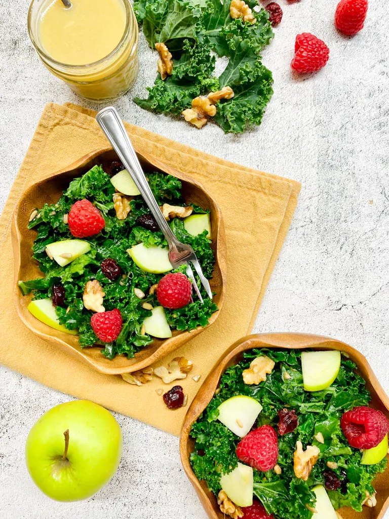 2 bowls of kale apple salad in wood bowls with a jar or dressing and a green apply on the side