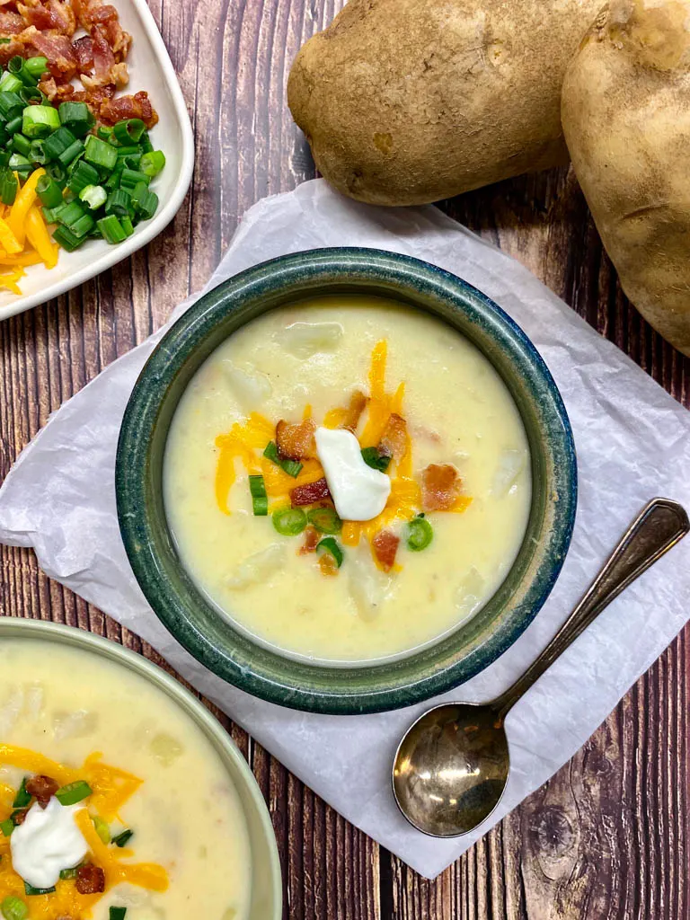 two bowls of loaded baked potato soup on a wood background with two whole potatoes and a spoon