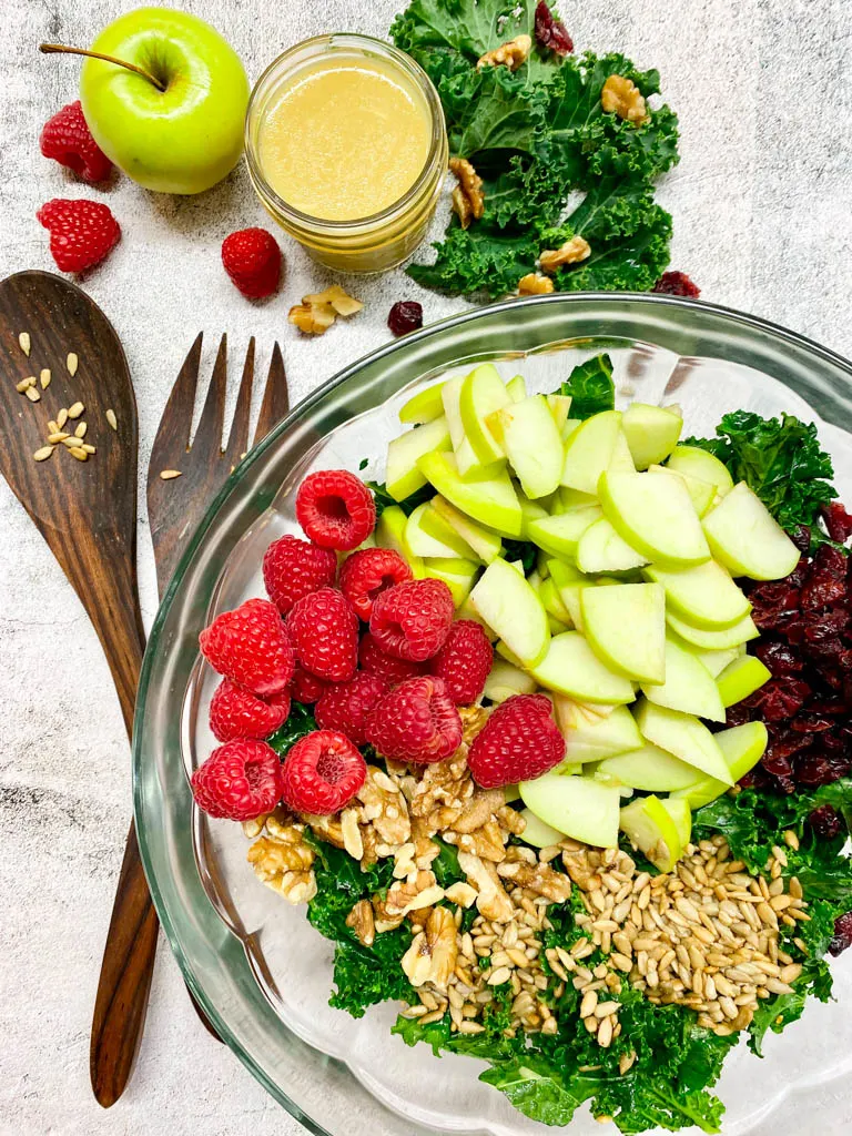 bowl of fall kale apple salad with an orange towel, wood spoons, salad dressing and green apple