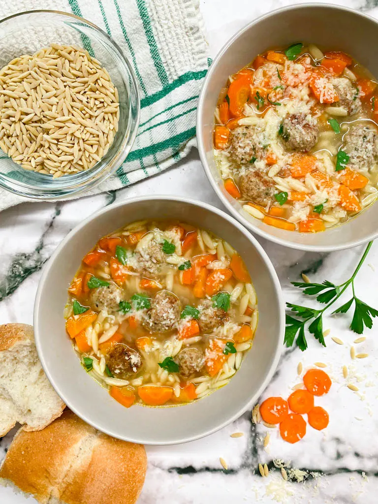 2 bowls of turkey meatball orzo soup with bread, carrots, onion, garlic, parsley and parmesan cheese around the bowls