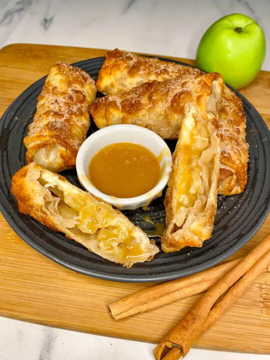 caramel apple cheesecake egg rolls on a black plate with a green apple