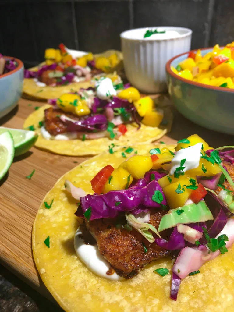 fish tacos plated on a wood board with line, mango salsa, cabbage slaw and lime crema