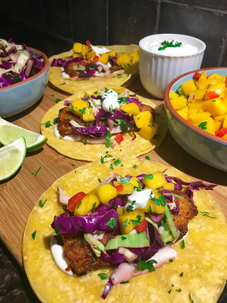 fish tacos plated on a wood board with line, mango salsa, cabbage slaw and lime crema