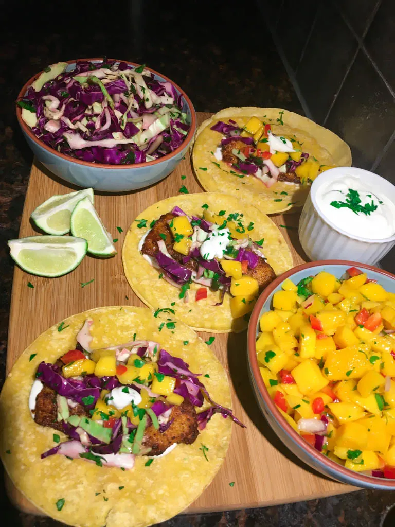 fish tacos plated on a wood board with line, mango salsa, cabbage slaw and lime crema