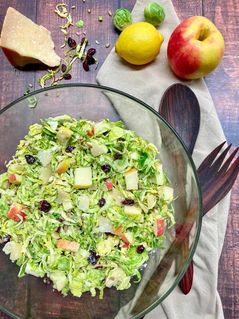 shaved brussels sprout salad in a glass bowl with apple, lemon, parmesan cheese and serving utensils on the side