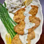 chicken costoletta on a beige plate with lemon gravy, asparagus, mashed potatoes and lemon wedge