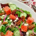 watermelon salad with feta cheese, candied pecans and red onion on a white platter