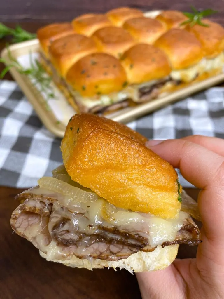 holding a roast beef slider with cheese and caramelized onions on a sheet pan garnished with rosemary