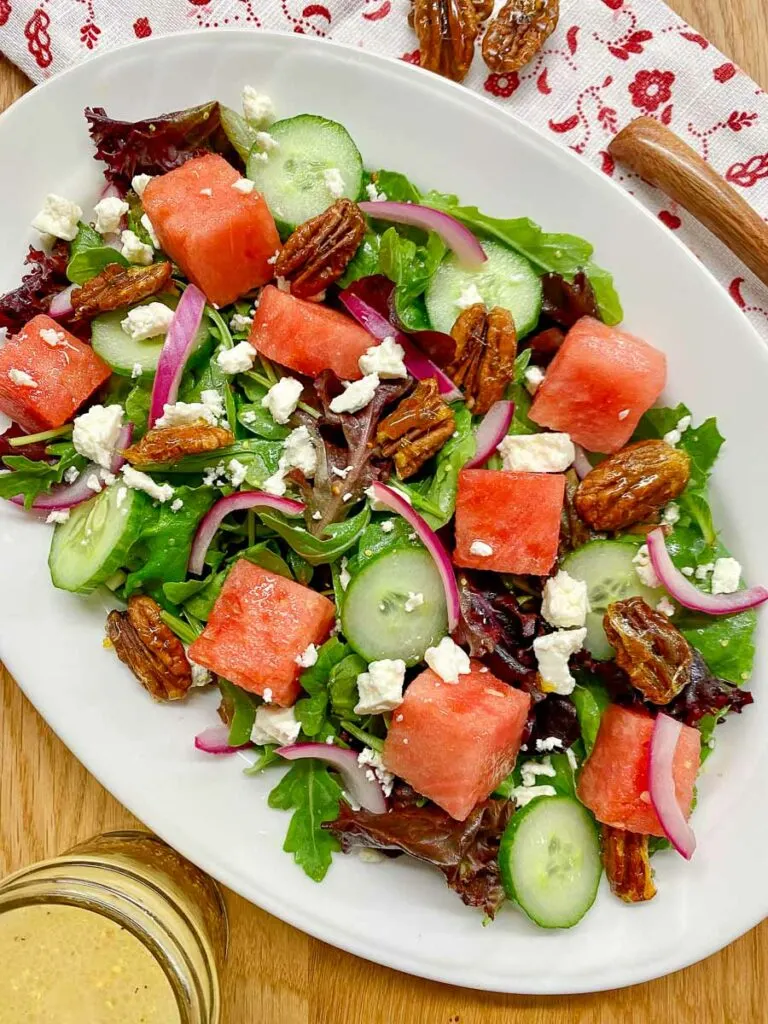 watermelon salad with feta cheese, candied pecans and red onion on a white platter