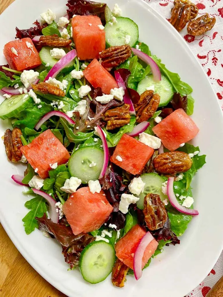 watermelon salad with feta cheese, candied pecans and red onion on a white platter