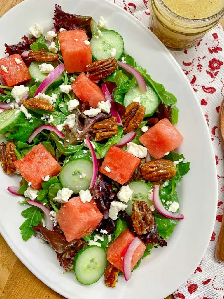 watermelon salad with feta cheese, candied pecans and red onion on a white platter