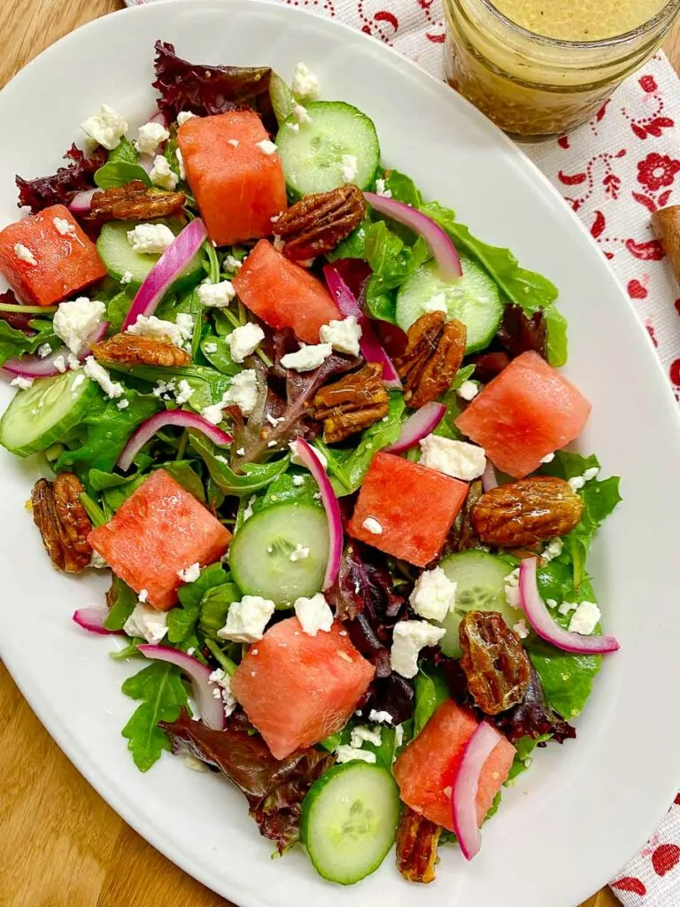 watermelon salad with feta cheese, candied pecans and red onion on a white platter