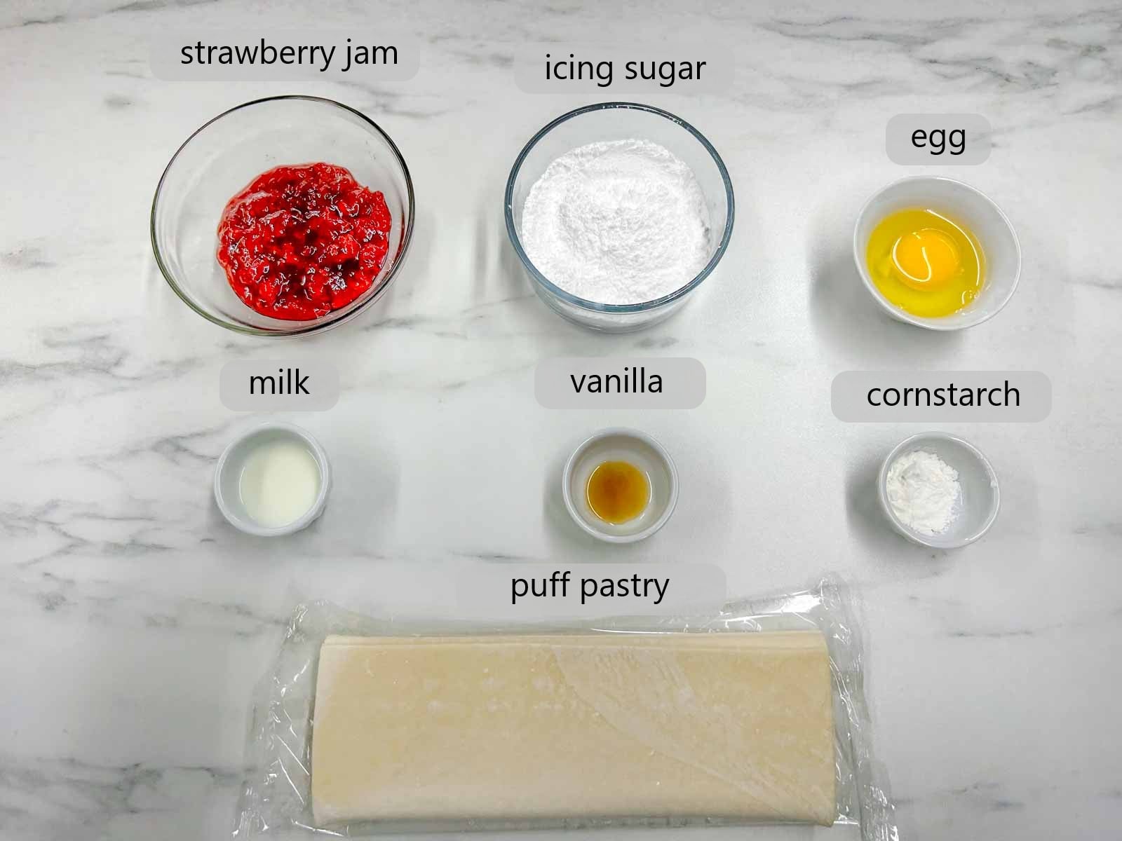 kitchen counter with bowls of ingredients and text overlays reading strawberry jam, icing sugar, egg, milk, vanilla, cornstarch, and puff pastry.
