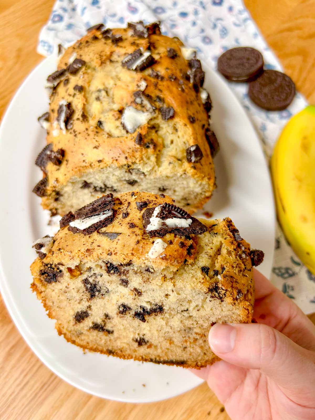 a loaf of Oreo banana bread is on a white serving platter. one piece of bread is held up close to the camera.
