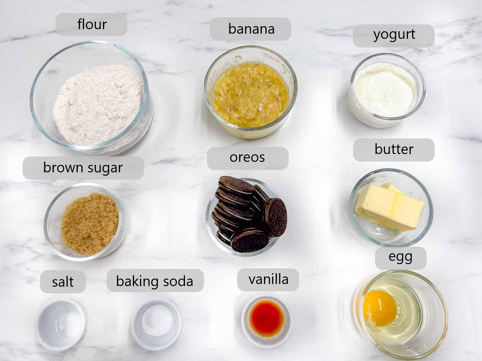 bowls with the ingredients to make Oreo banana bread are on a white kitchen counter with text labels reading - flour, banana, yogurt, brown sugar, Oreos, butter, salt, baking soda, vanilla, and egg.