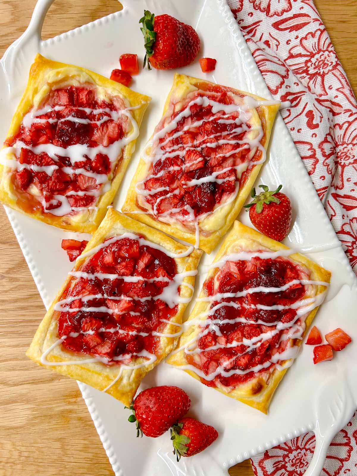 a white platter has strawberry cheese Danish on top that are drizzled with icing. there are additional strawberries on the plate for garnish.