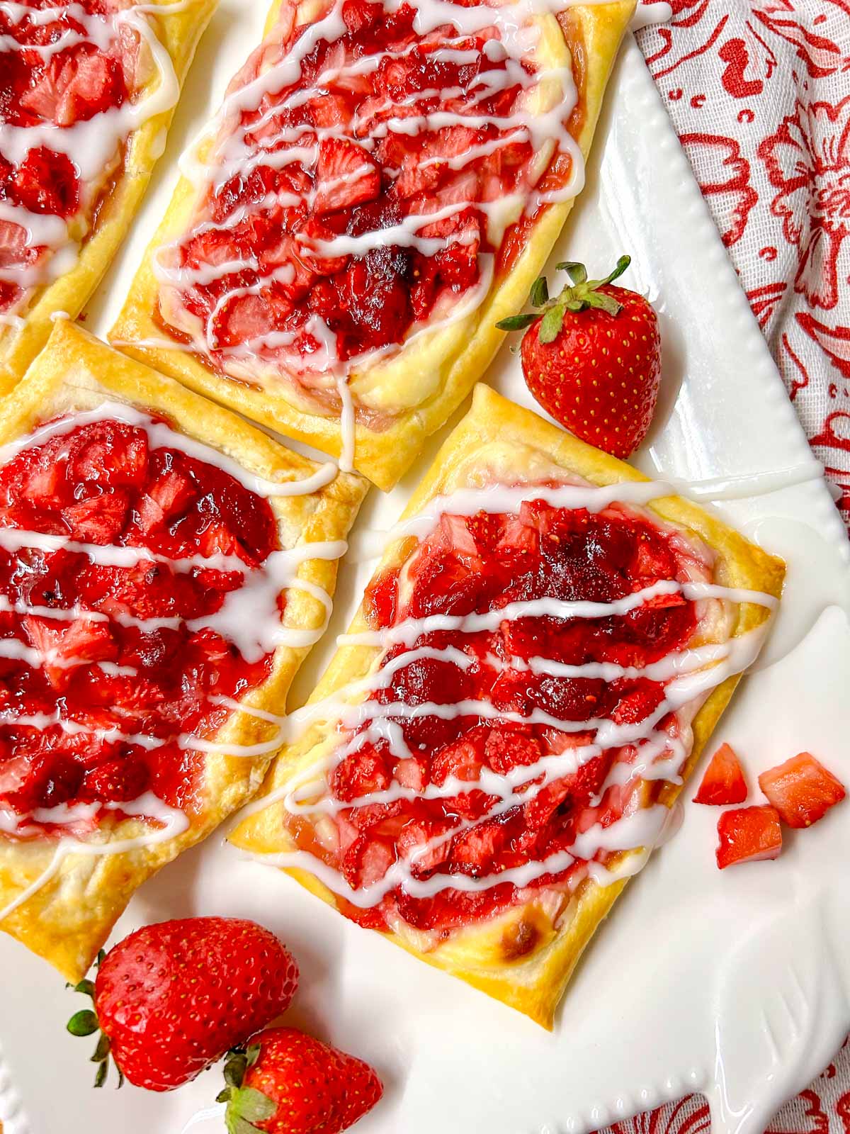 a white platter has strawberry cheese Danish on top that are drizzled with icing. there are additional strawberries on the plate for garnish.