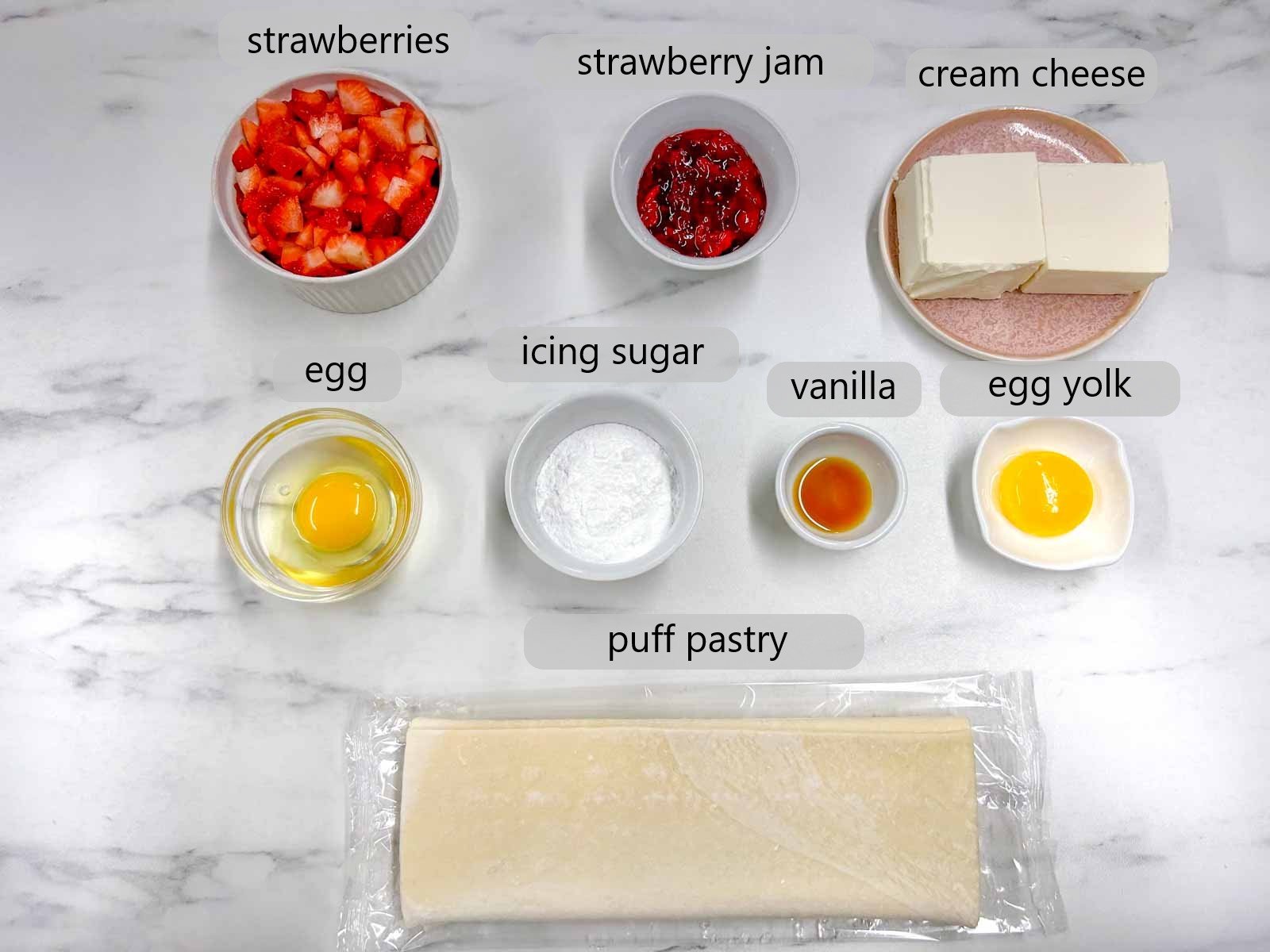 a white marble counter top has ingredients in them with text overlay labels reading strawberries, strawberry jam, cream cheese, egg, icing sugar, vanilla, egg yolk, and puff pastry.