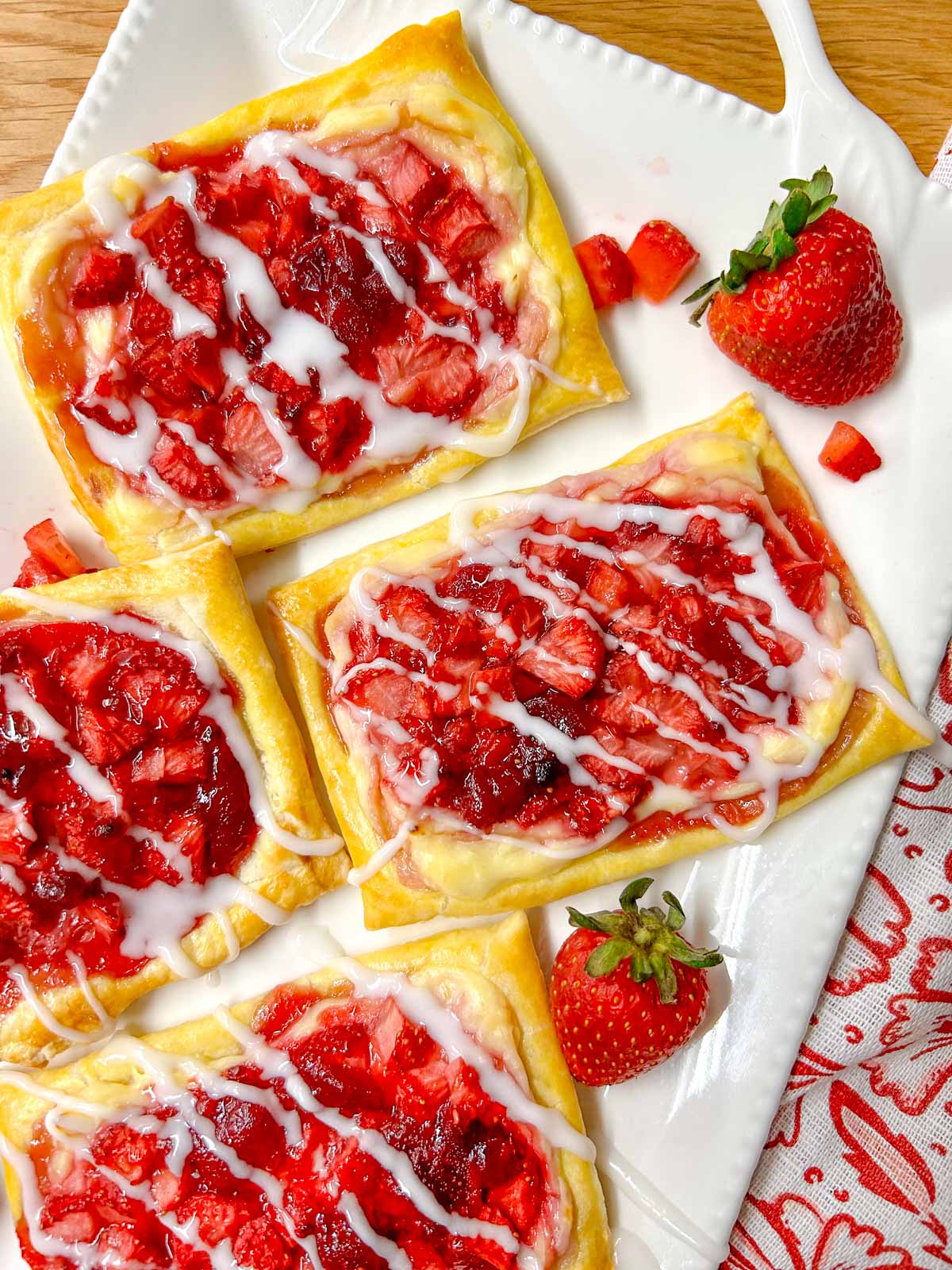 a white platter has strawberry cheese Danish on top that are drizzled with icing. there are additional strawberries on the plate for garnish.