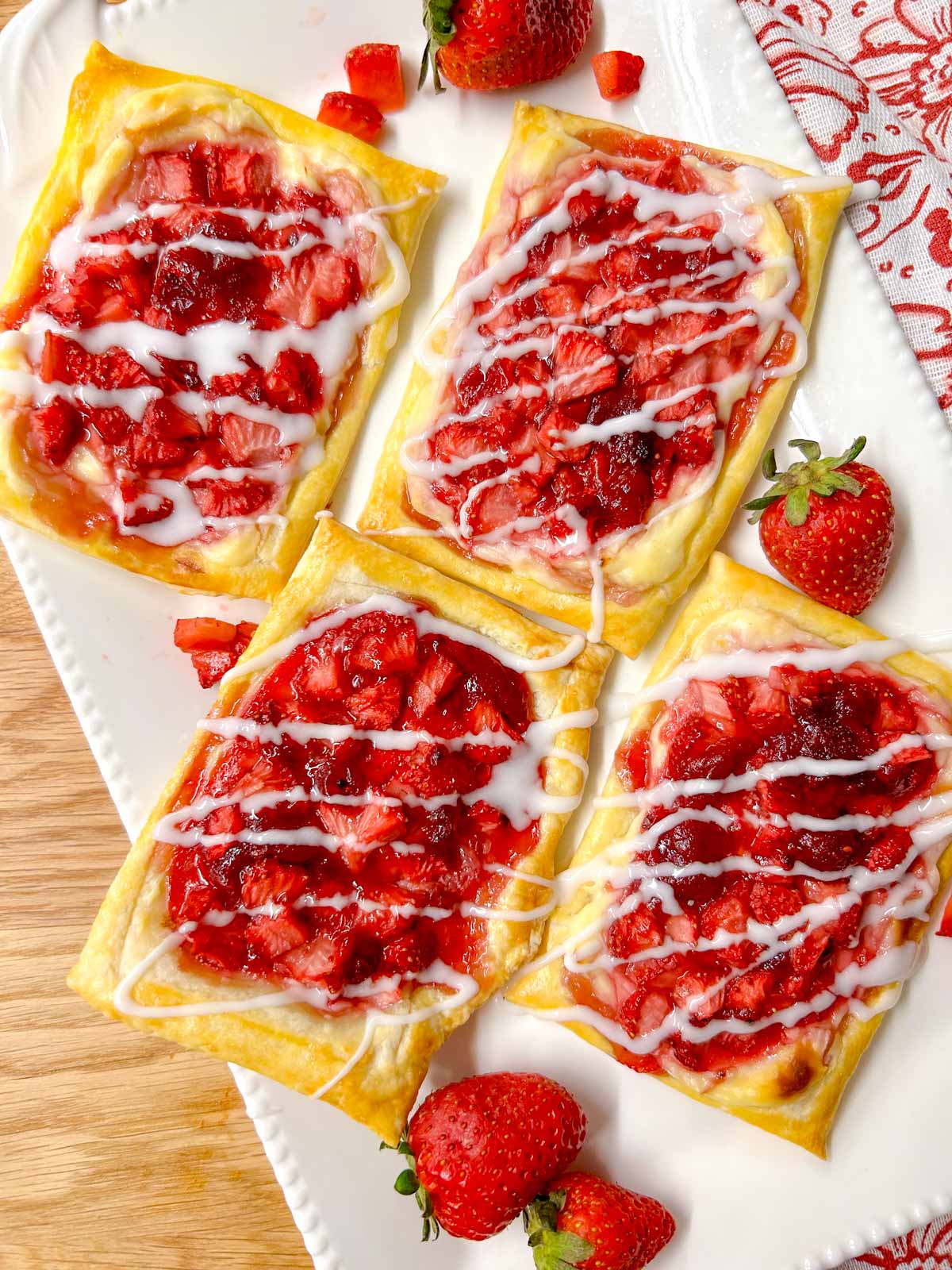 a white platter has strawberry cheese Danish on top that are drizzled with icing. there are additional strawberries on the plate for garnish.