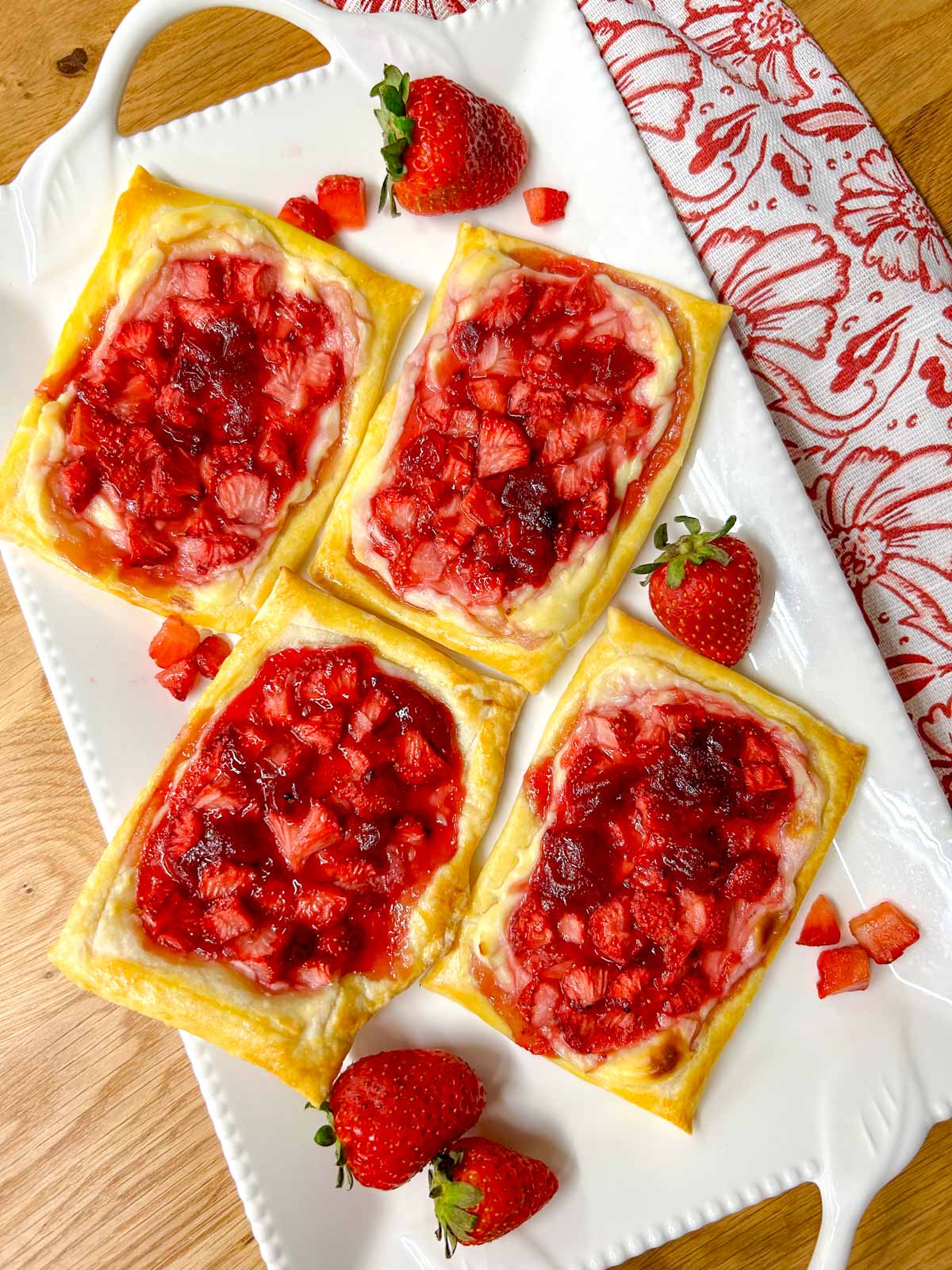 a white platter has strawberry cheese Danish on top that are drizzled with icing. there are additional strawberries on the plate for garnish.