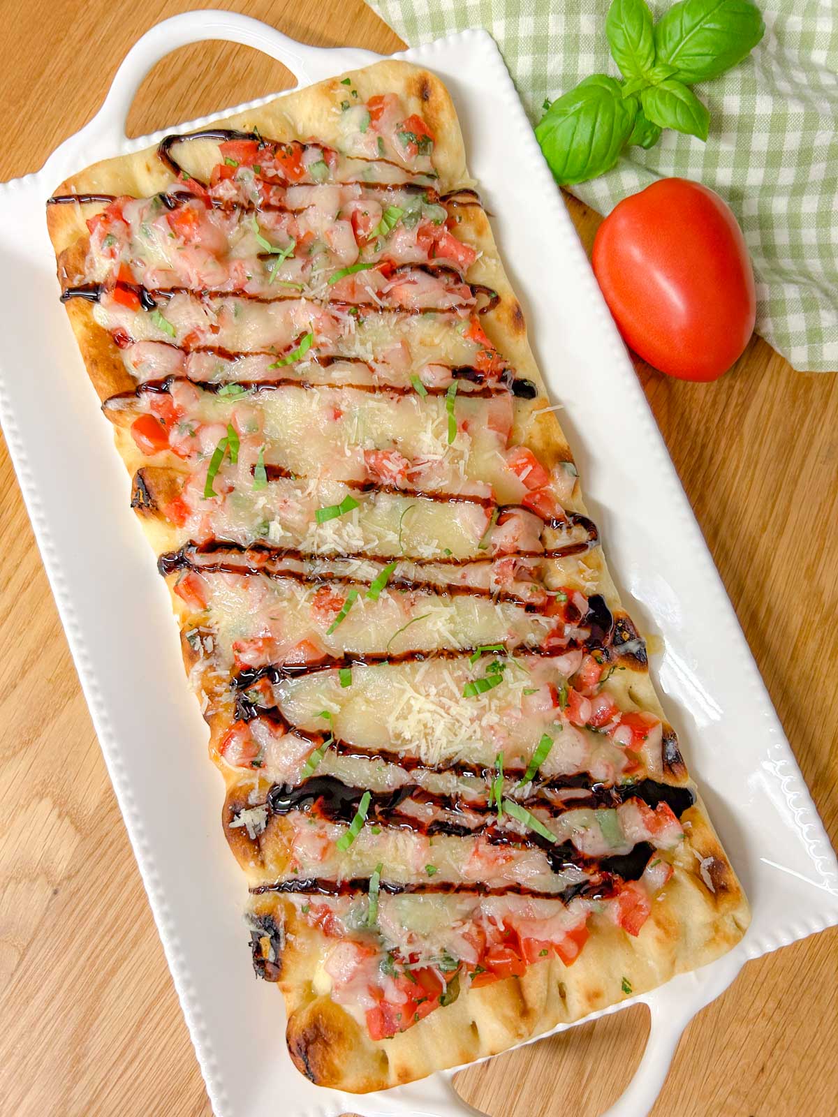 a bruschetta flatbread pizza drizzled with balsamic glaze and garnished with basil is on a white serving platter.