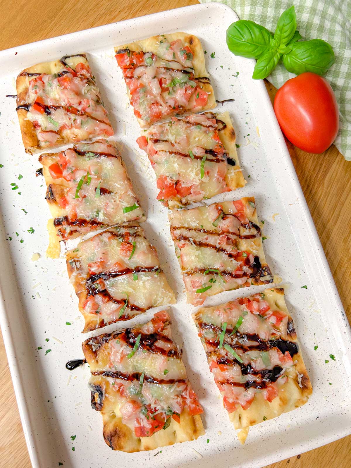a bruschetta flatbread pizza drizzled with balsamic glaze and garnished with basil is on a white serving platter.