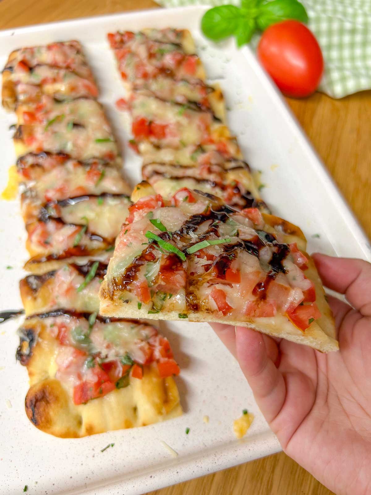 a cut up bruschetta flatbread pizza drizzled with balsamic glaze and garnished with basil is on a white serving platter. one piece of flatbread is help up close to the camera.