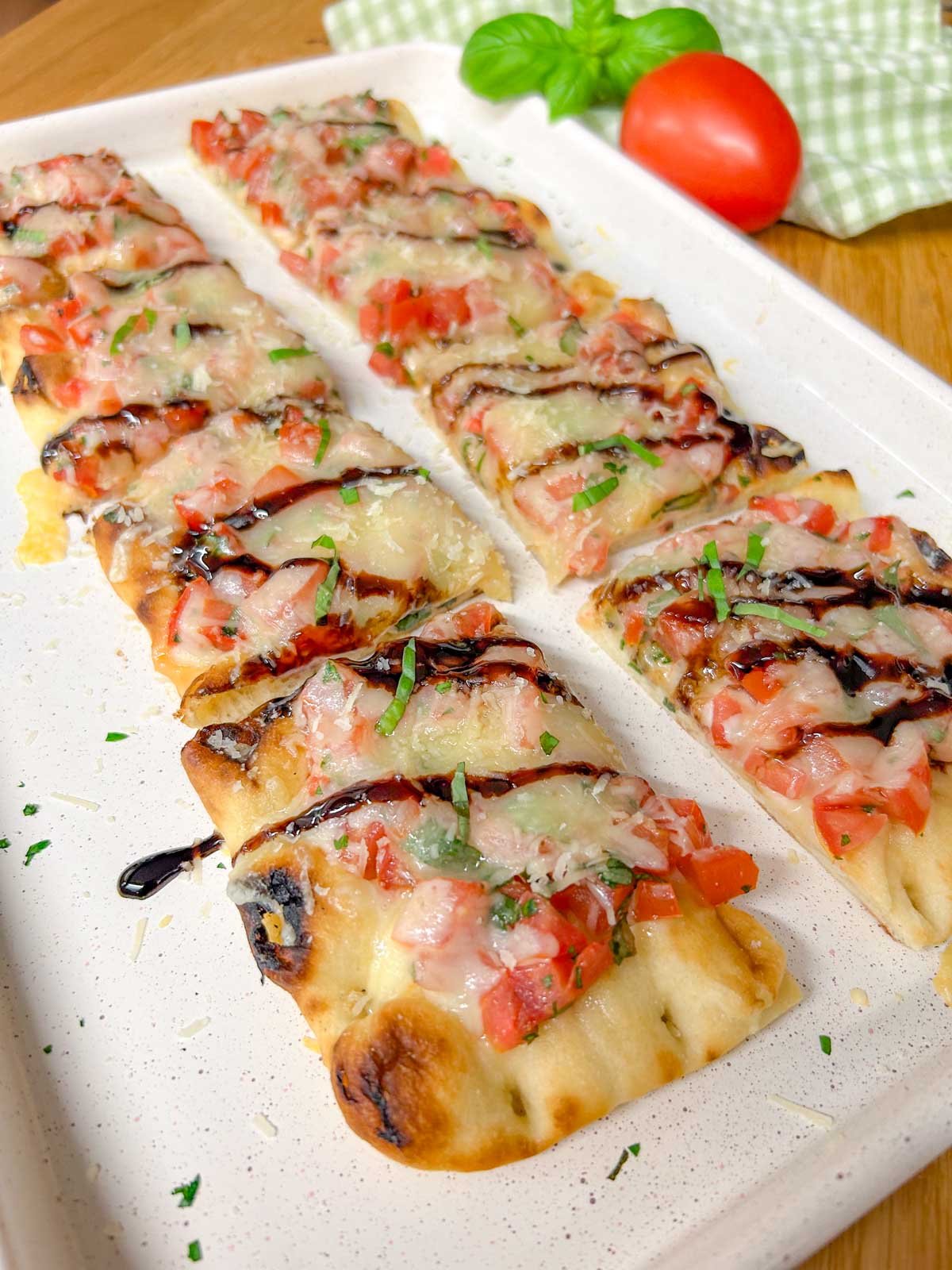 a cut up bruschetta flatbread pizza drizzled with balsamic glaze and garnished with basil is on a white serving platter.