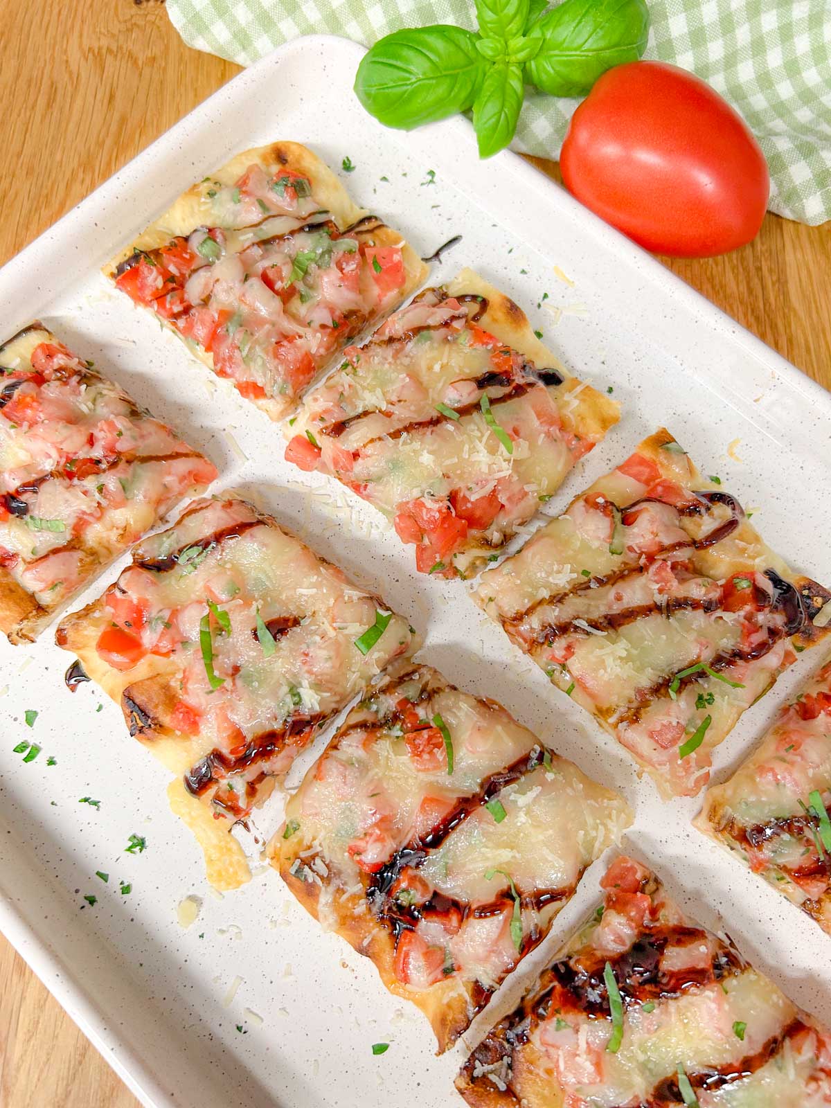 a cut up bruschetta flatbread pizza drizzled with balsamic glaze and garnished with basil is on a white serving platter.