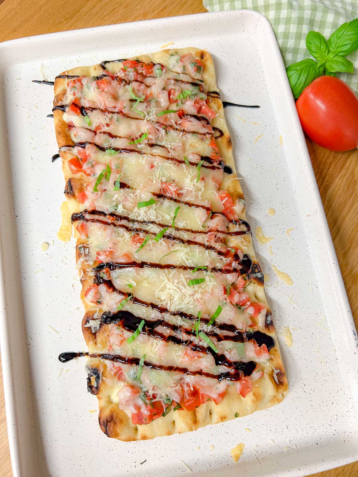 a bruschetta flatbread pizza drizzled with balsamic glaze and garnished with basil is on a white serving platter.