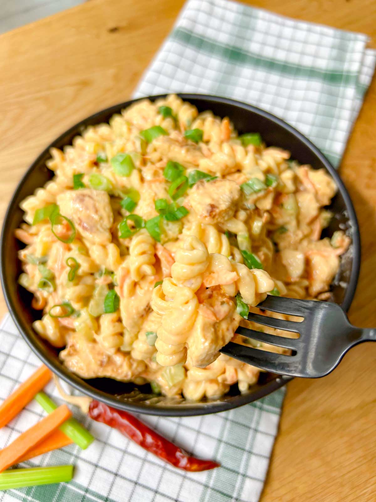 a black serving bowl is filled with buffalo chicken pasta salad and garnished with sliced green onions. a black fork is holding a bite of the salad.