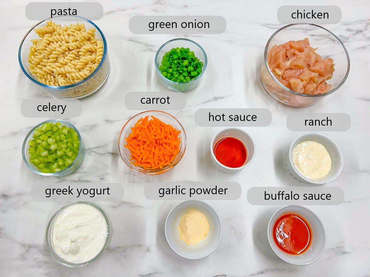 clear glass bowl with labels for the ingredients, pasta, green onion, chicken, celery, carrot, hot sauce, ranch, greek yogurt, garlic powder and buffalo sauce, on a white marble counter top