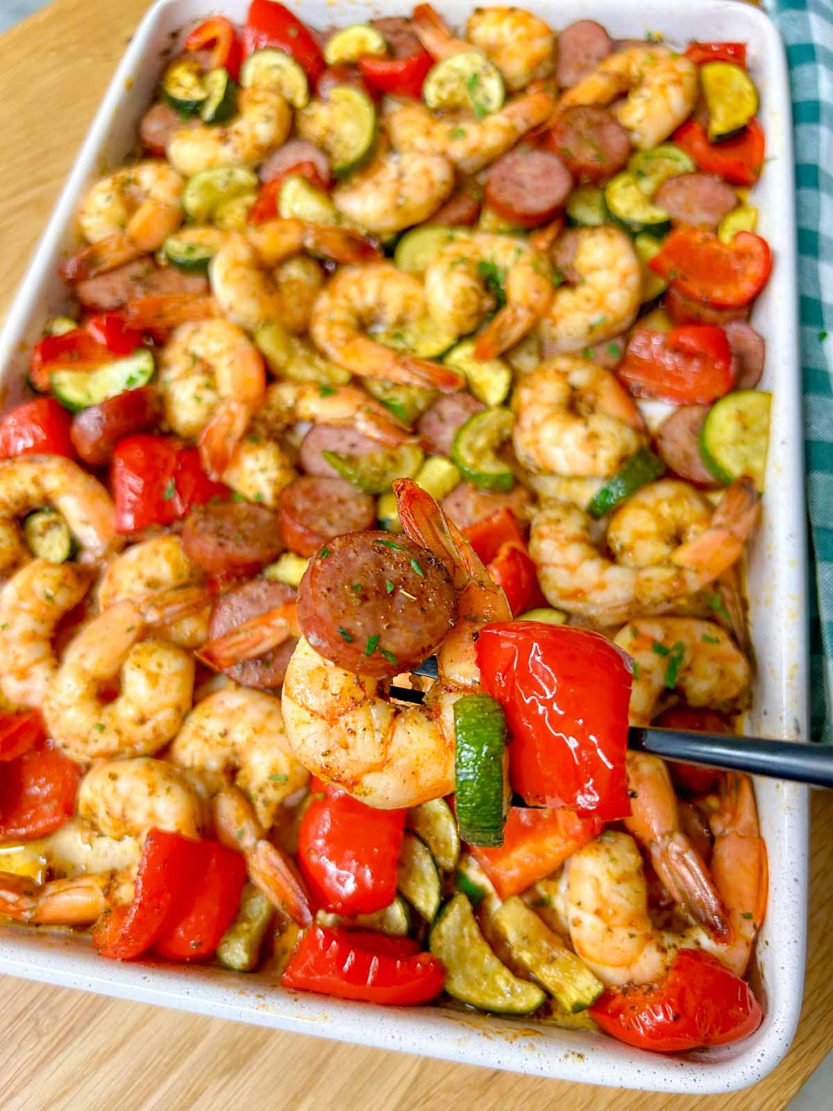 a white sheet pan is filled with a Cajun shrimp and sausage sheet pan bake on a wood counter top. a black fork is holding a shrimp and pepper.