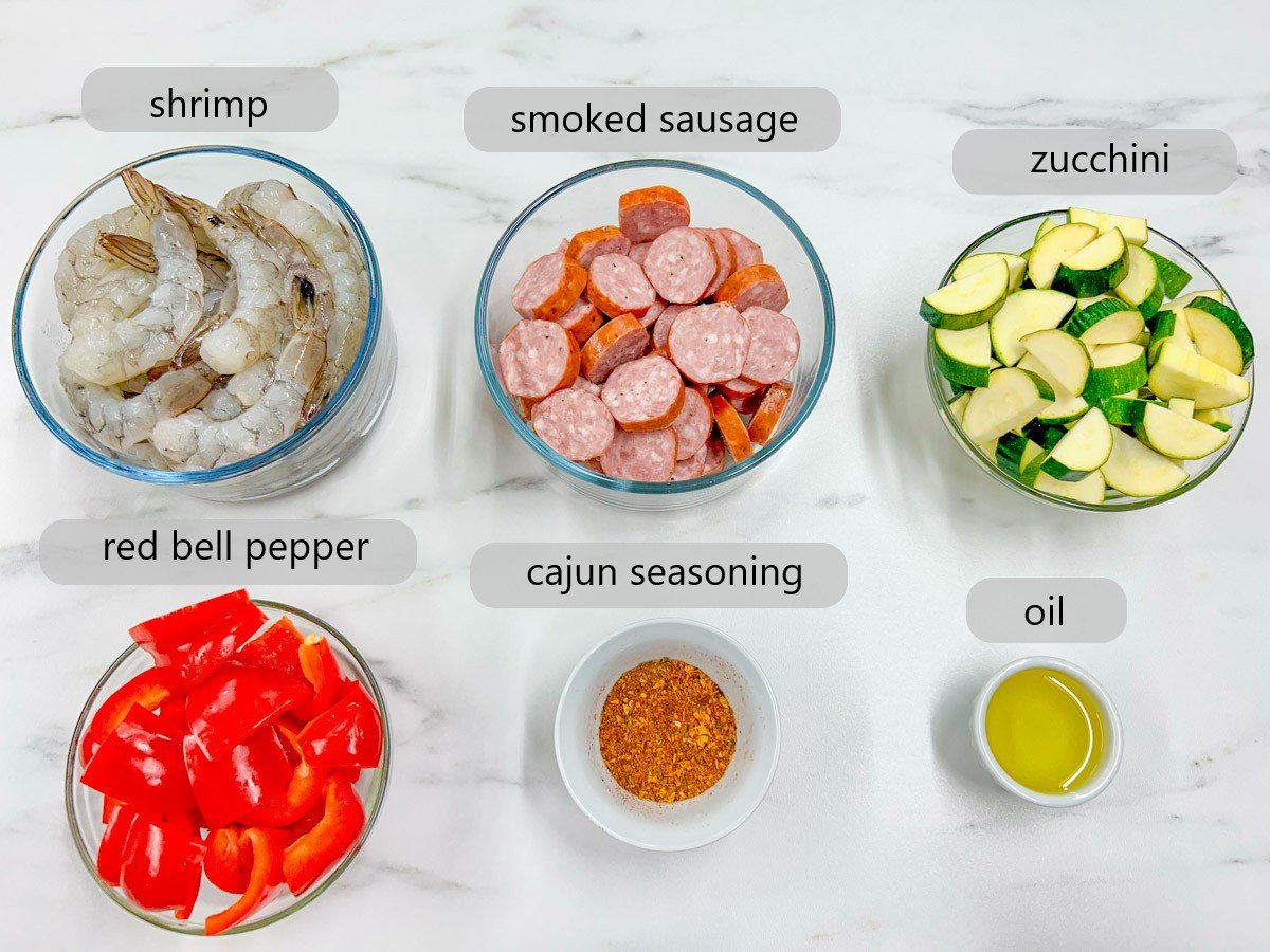 a white marble counter top has bowls with the ingredients to make a Cajun shrimp and sausage sheet pan with text overlay labels that read - shrimp, smoked sausage, zucchini, red bell pepper, Cajun seasoning and oil.
