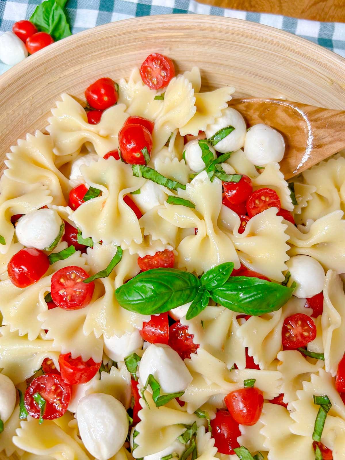 a wooden bowl is filled with caprese pasta salad and garnished with fresh basil. a wooden spoon is holding some of the pasta