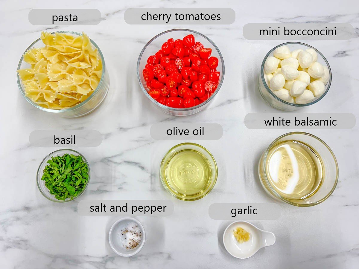 bowls with the ingredients and labels above them on a white marble counter top. pasta, cherry tomatoes, mini bocconcini, basil, olive oil, white balsamic, salt and pepper and garlic are shown in bowls.