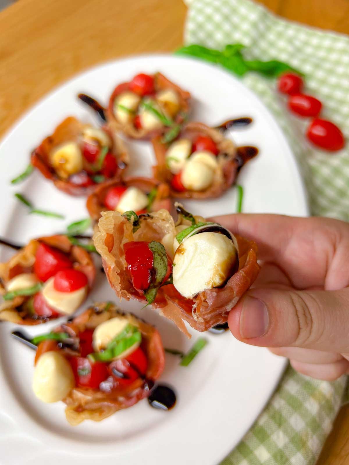 a white platter has crispy caprese prosciutto cups with balsamic glaze drizzled over top. one cup has a bite taken out and is held close to the camera.