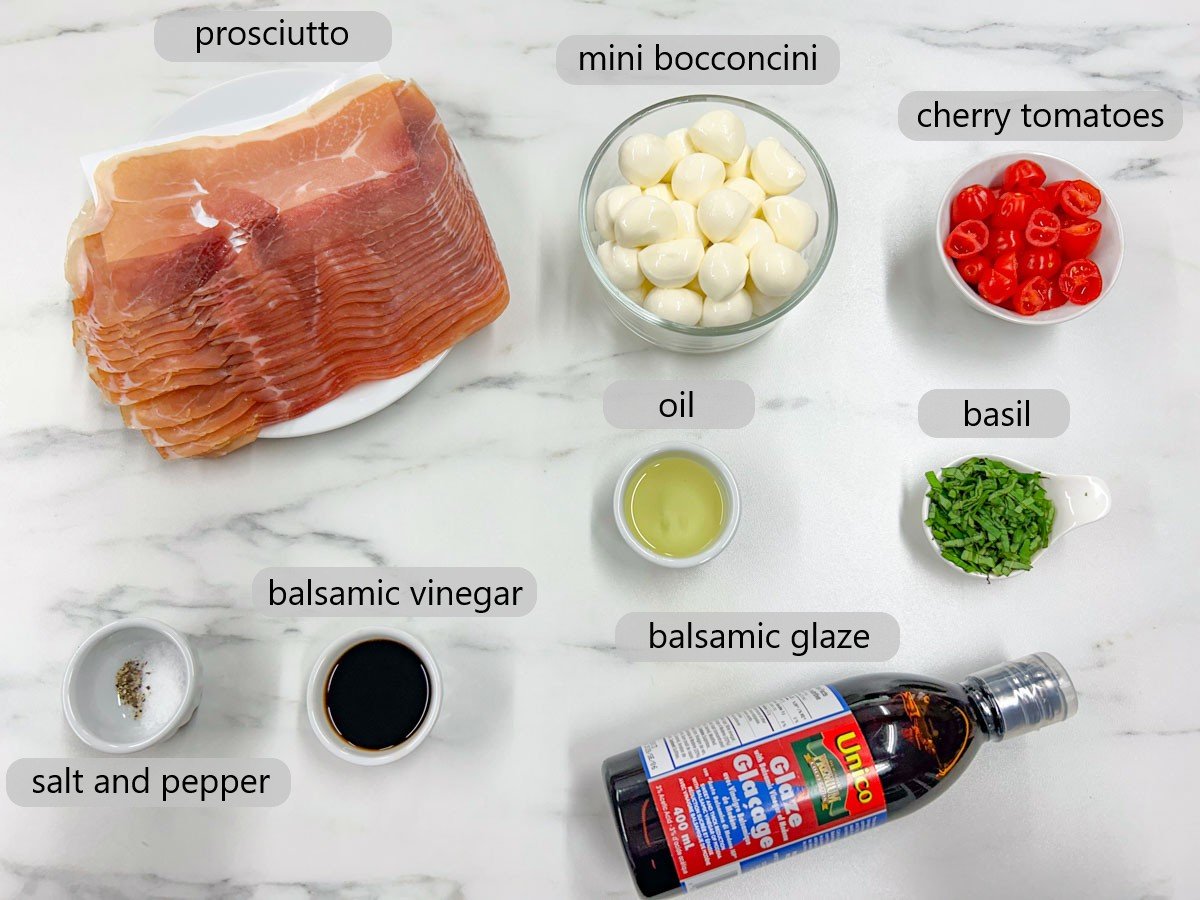 a white marble counter top has bowls with ingredients and text overlay reading prosciutto, mini bocconcini, cherry tomatoes, salt and pepper, balsamic vinegar, oil, basil, and balsamic glaze.