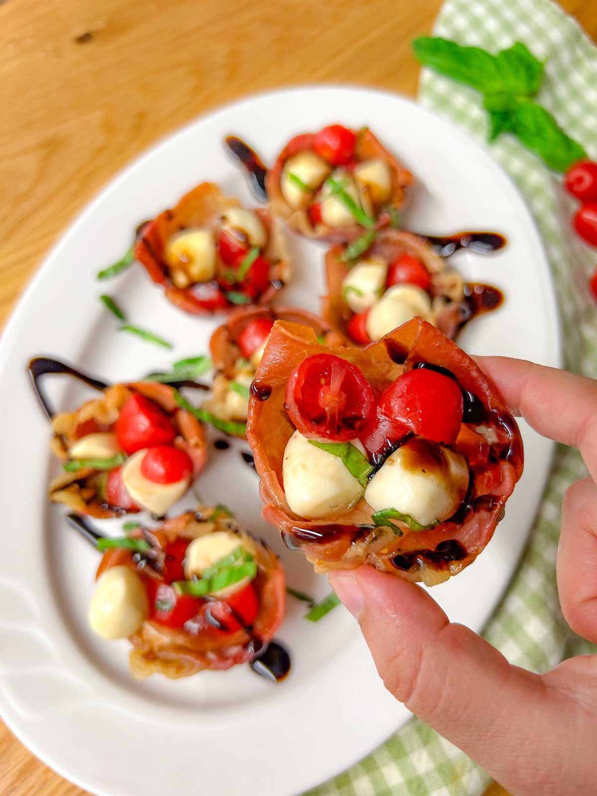 a white platter has crispy caprese prosciutto cups with balsamic glaze drizzled over top. one cup is held closer to the camera.