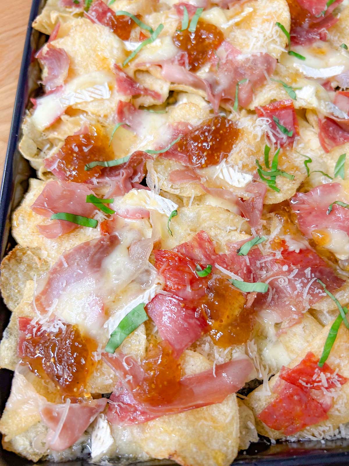 a black rimmed sheet pan is filled with baked charcuterie nachos and garnished with parmesan cheese and sliced basil.