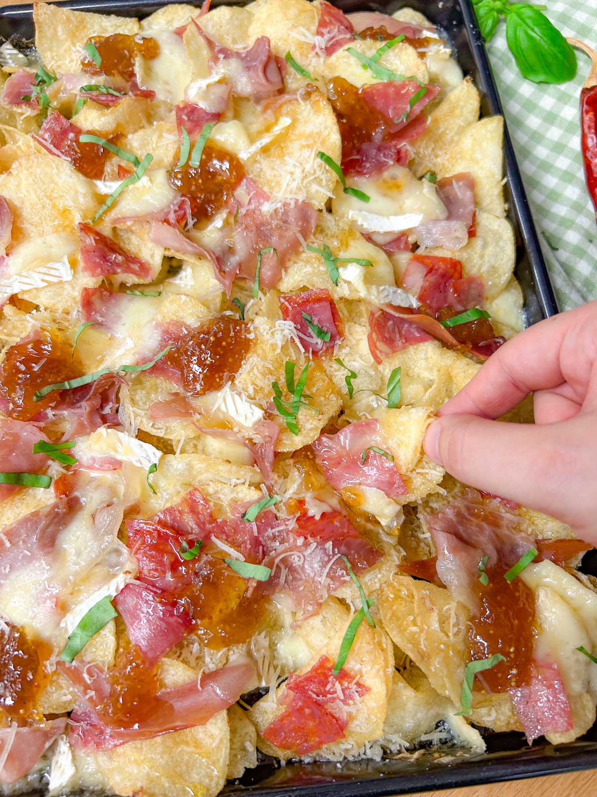 a black rimmed sheet pan is filled with baked charcuterie nachos and garnished with parmesan cheese and sliced basil. a hand if picking up one chip.