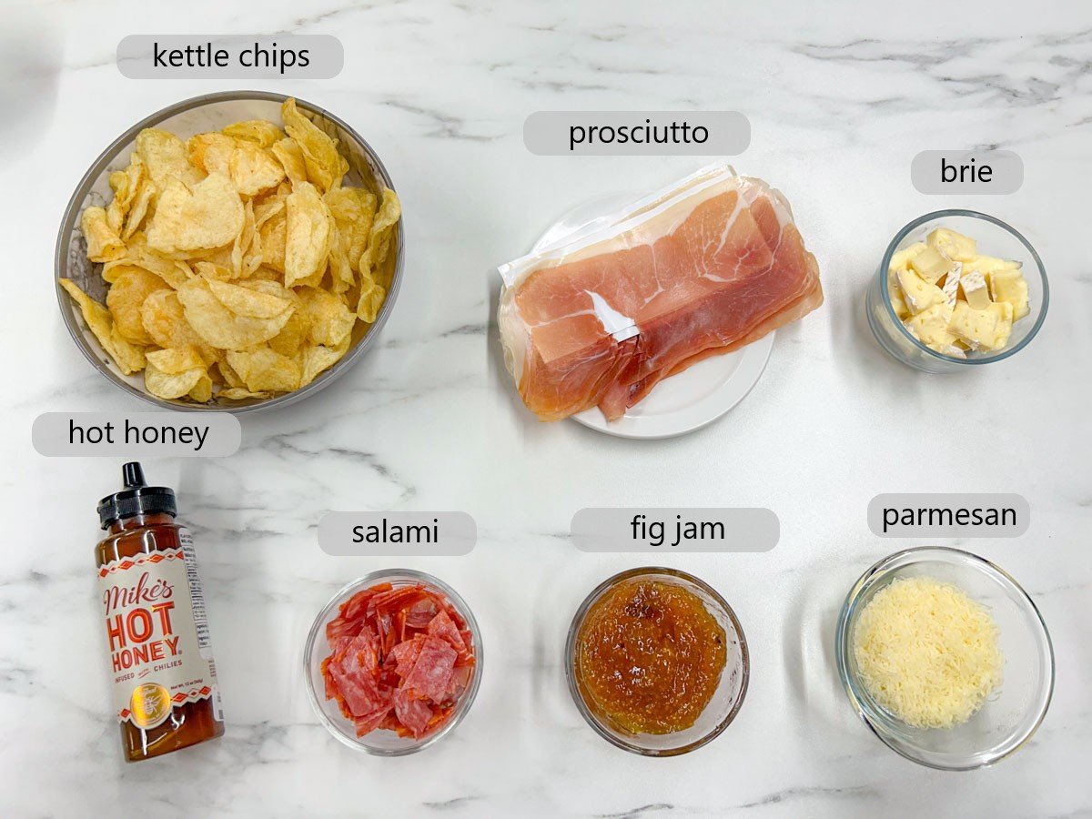 a white marble counter top has bowls with ingredients and labels reading kettle chips, prosciutto, brie, hot honey, salami, fig jam, and parmesan.