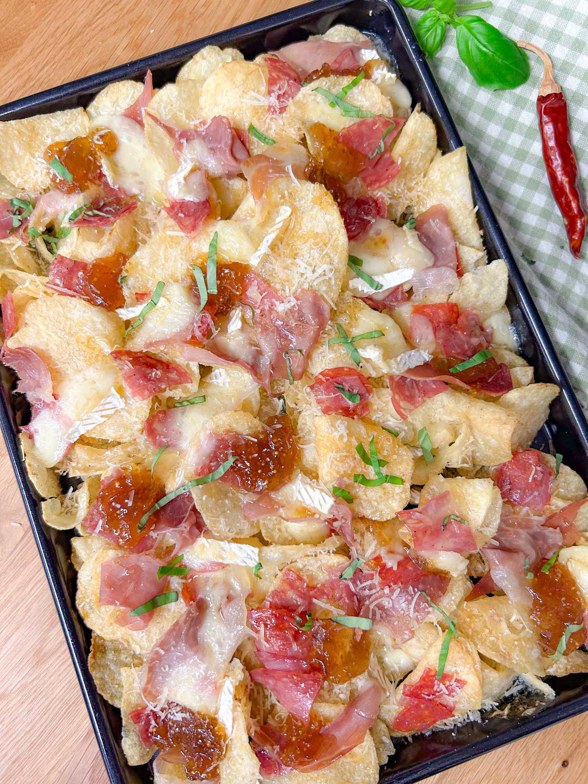 a black rimmed sheet pan is filled with baked charcuterie nachos and garnished with parmesan cheese and sliced basil.