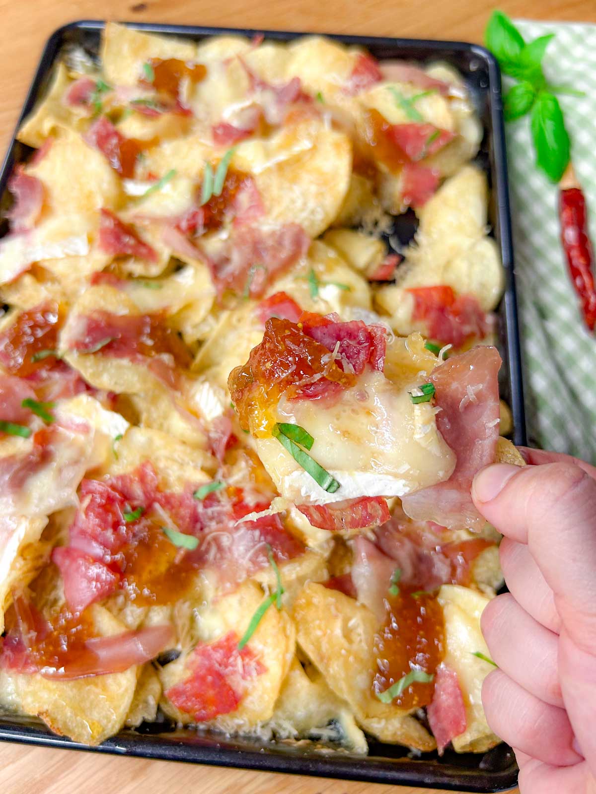 a black rimmed sheet pan is filled with baked charcuterie nachos and garnished with parmesan cheese and sliced basil. one chip is held closer to the camera.
