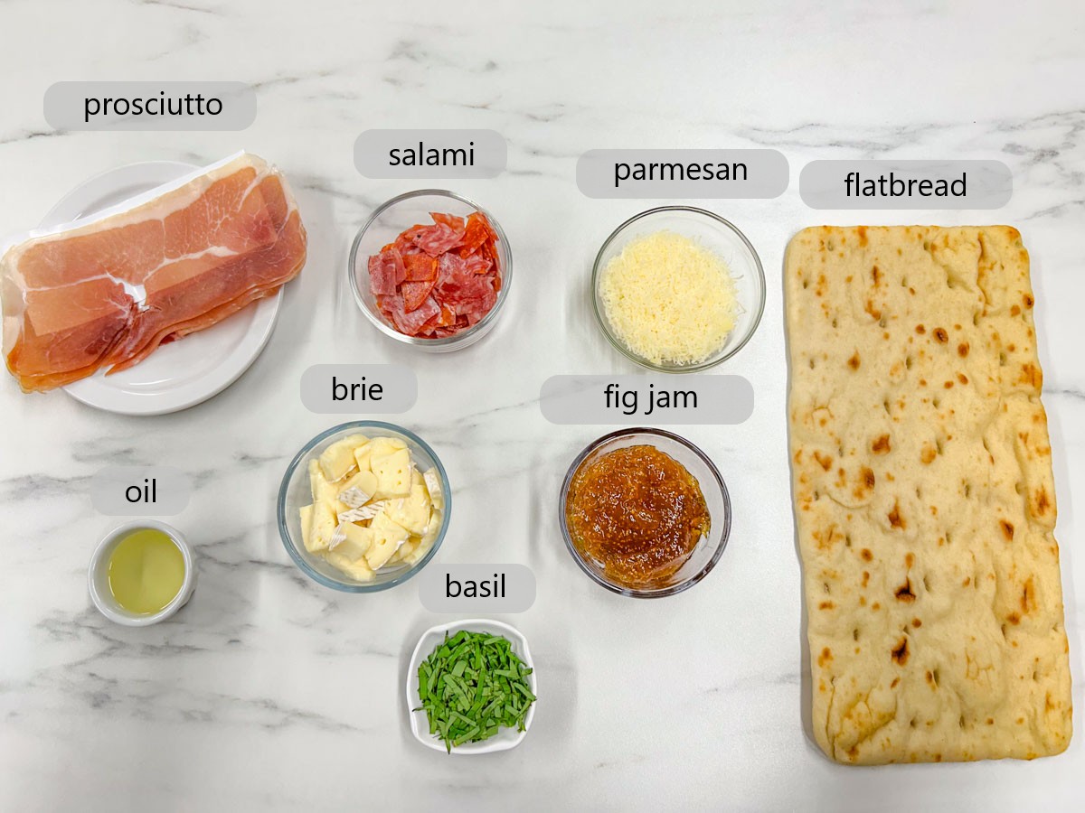 a white marble countertop has bowls with ingredients and text overlays reading prosciutto, salami, parmesan, flatbread, oil, brie, fig jam, and basil.