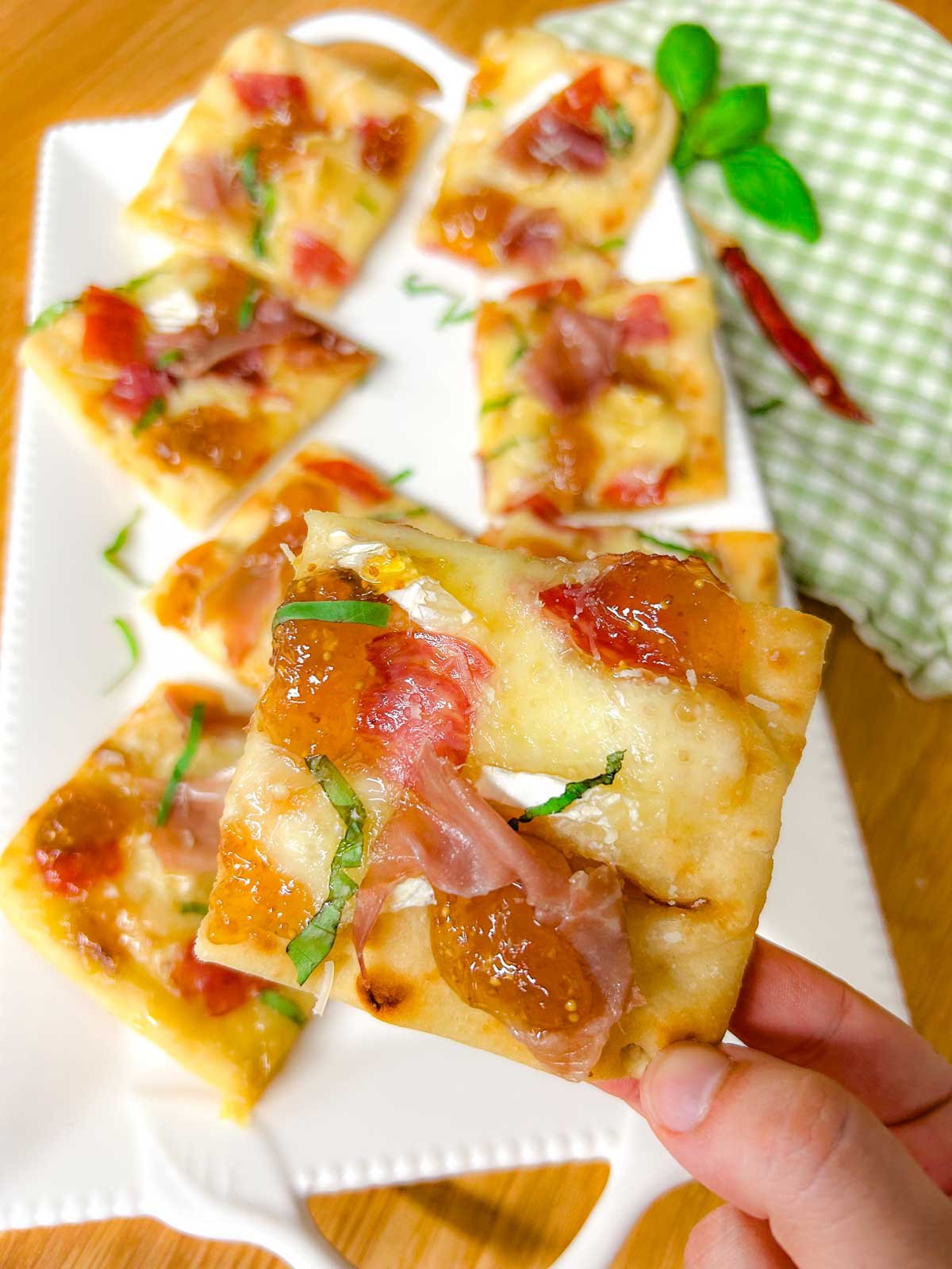 a white platter has a charcuterie flatbread on top that is garnished with thinly sliced basil. one cut piece is held up closer to the camera.