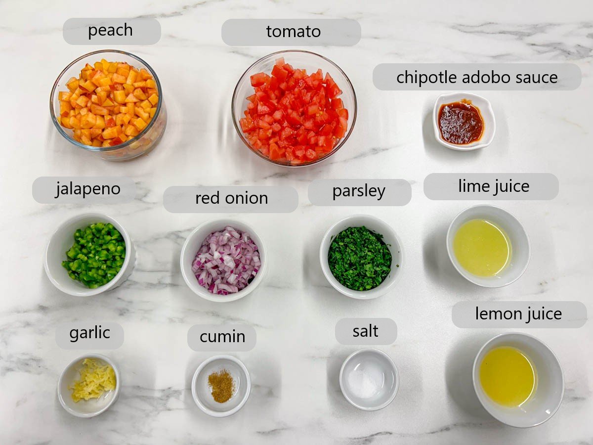 a white marble counter top as bowl with the ingredients labelled as peach, tomato, chipotle adobo sauce, jalapeno, red onion, parsley, lime juice, garlic, cumin, salt, and lemon juice.