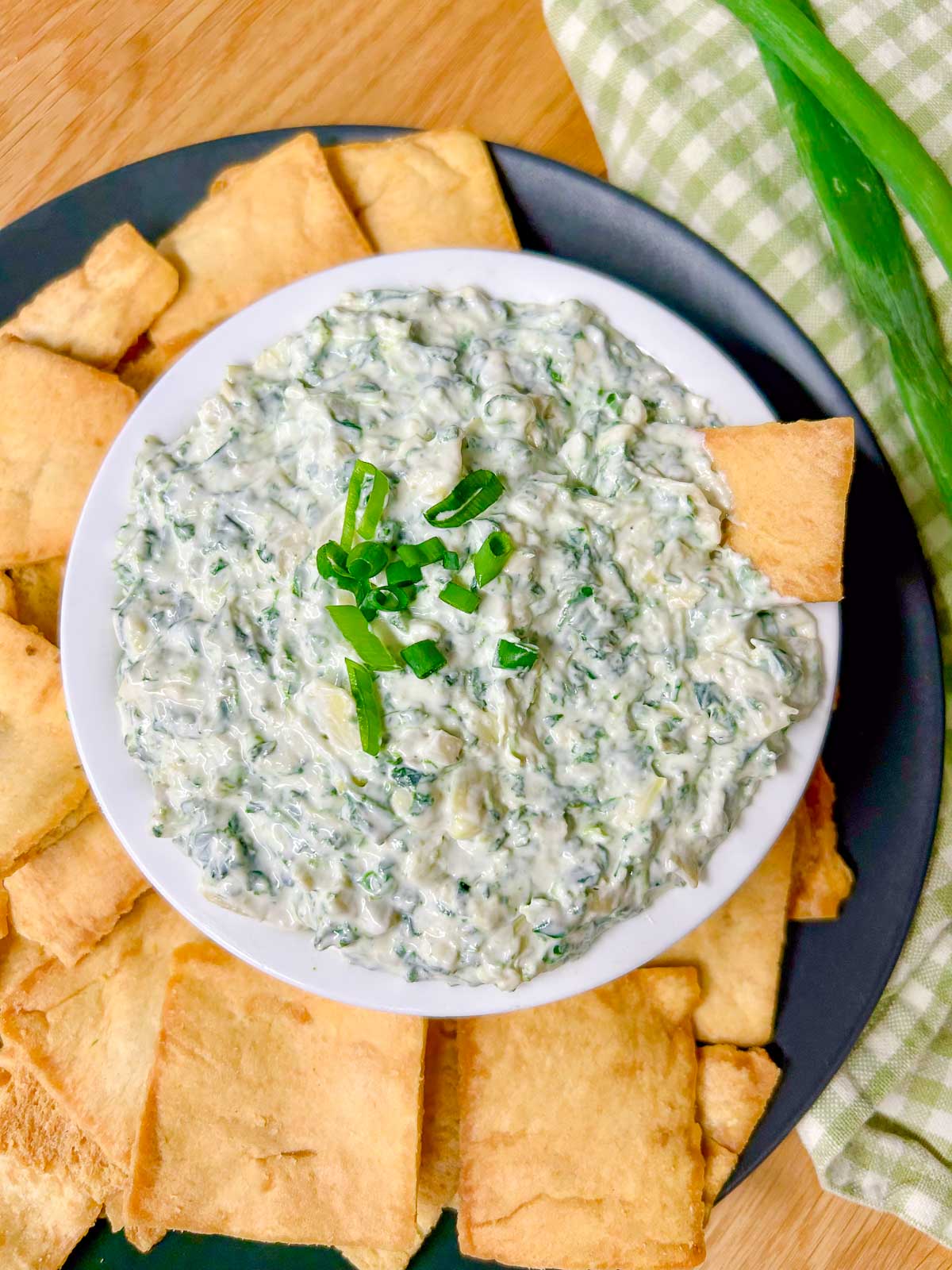 a black plate has a white serving bowl with spinach artichoke dip in it and pita chips around the bowl.